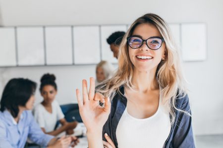 Close-up portrait of pretty female manager from sales department. Indoor photo of smiling girl working in office with discussing people on background.